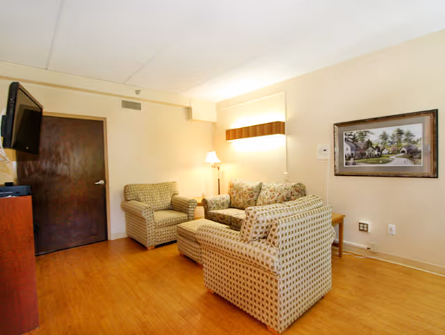 A cozy living room with wooden flooring, featuring a patterned armchair, a floral-patterned loveseat, and another patterned armchair arranged around a small wooden coffee table. A floor lamp and a wall-mounted light provide warm lighting. A framed picture hangs on the wall, and a dark wooden door is visible in the background. A flat-screen TV is mounted on the wall to the left.