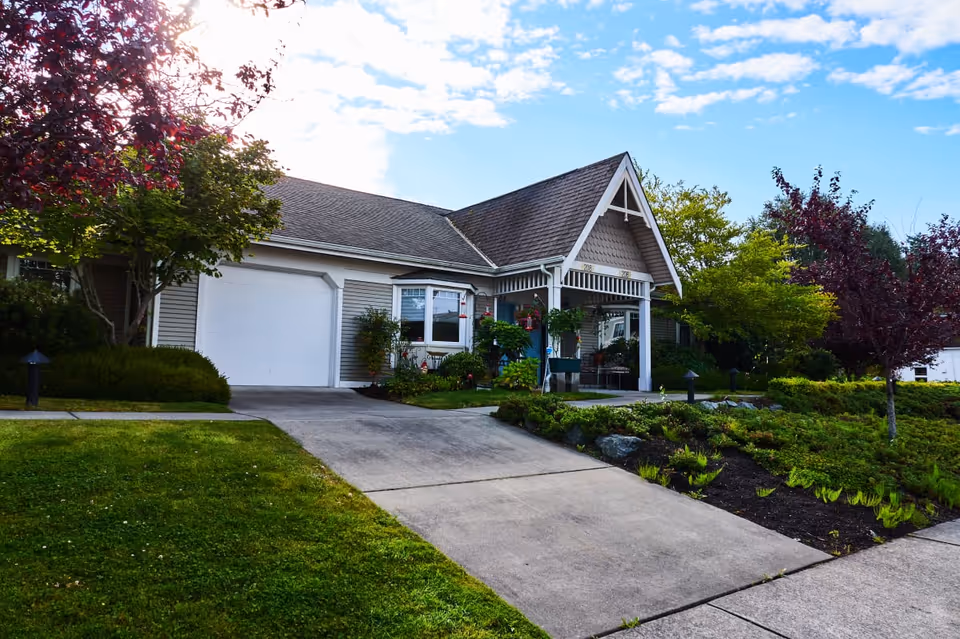 Exterior view of a single-story residential building with a sloped roof, a garage door, and a covered porch area surrounded by well-maintained greenery and trees under a partly cloudy sky.