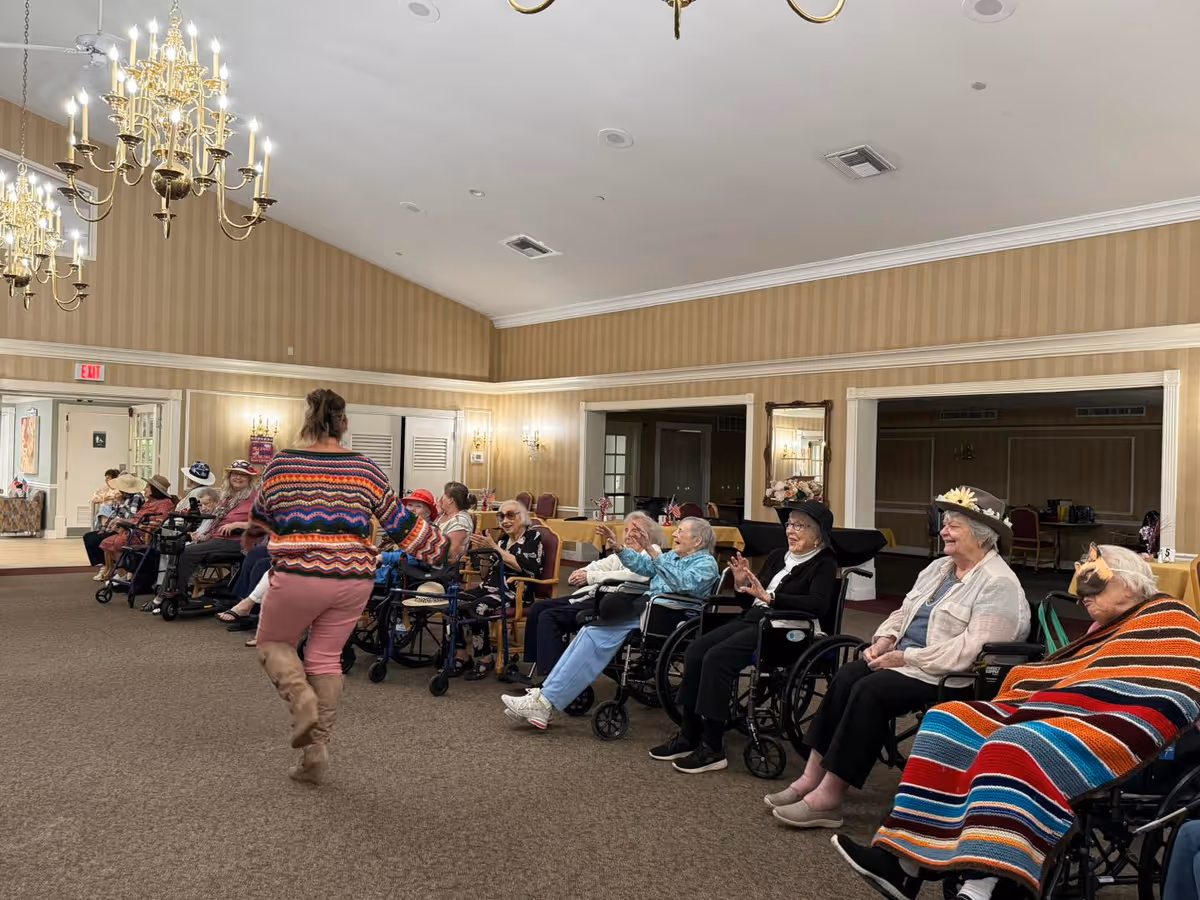 A group of elderly people, many in wheelchairs, seated in a spacious room with beige striped wallpaper and chandeliers. A woman in a colorful sweater and pink pants is walking in front of them, engaging with the group. Some of the elderly individuals are wearing hats, and one is covered with a colorful striped blanket.