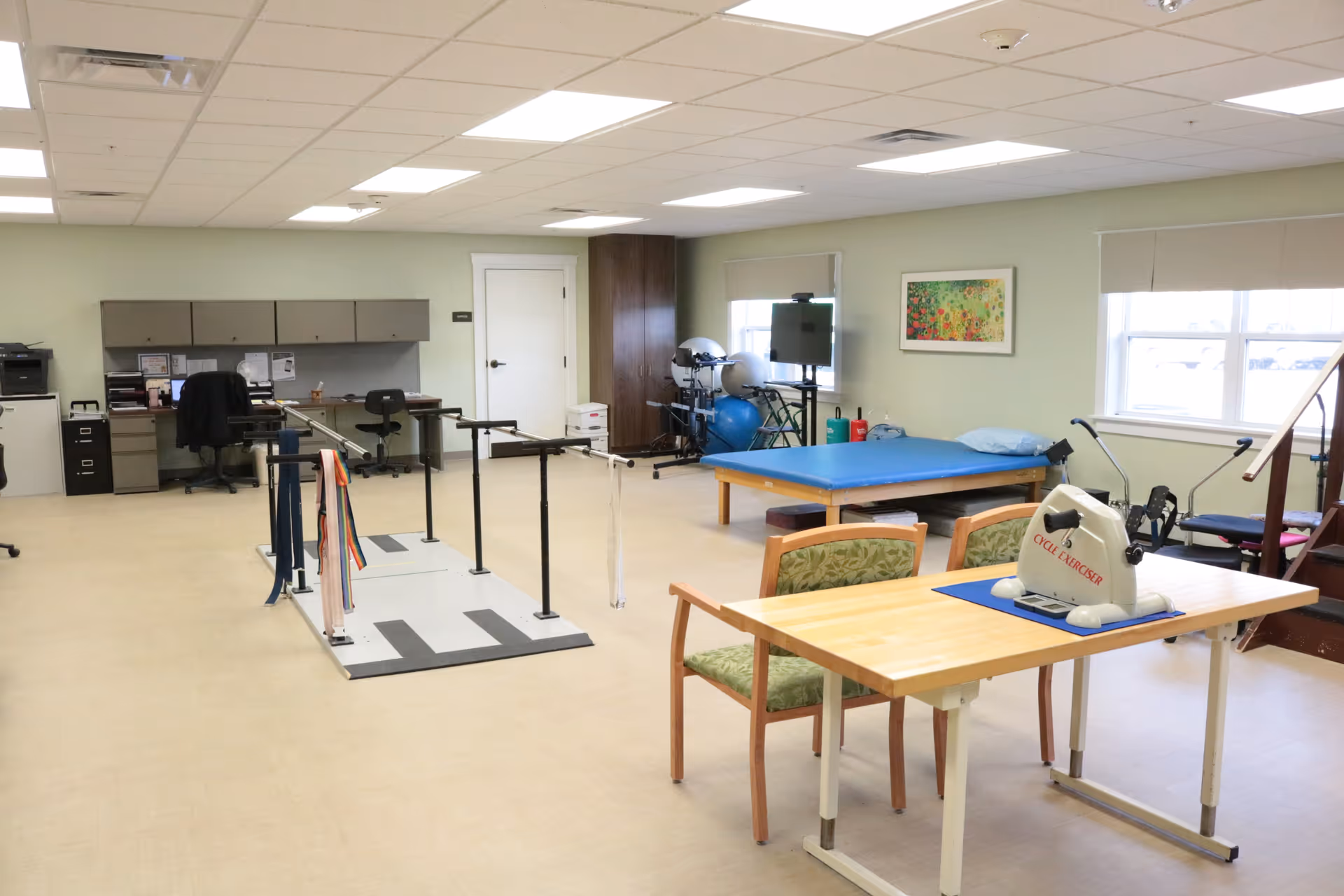 Spacious rehabilitation therapy room with parallel bars, an exercise table, chairs, desk area and fitness equipment.