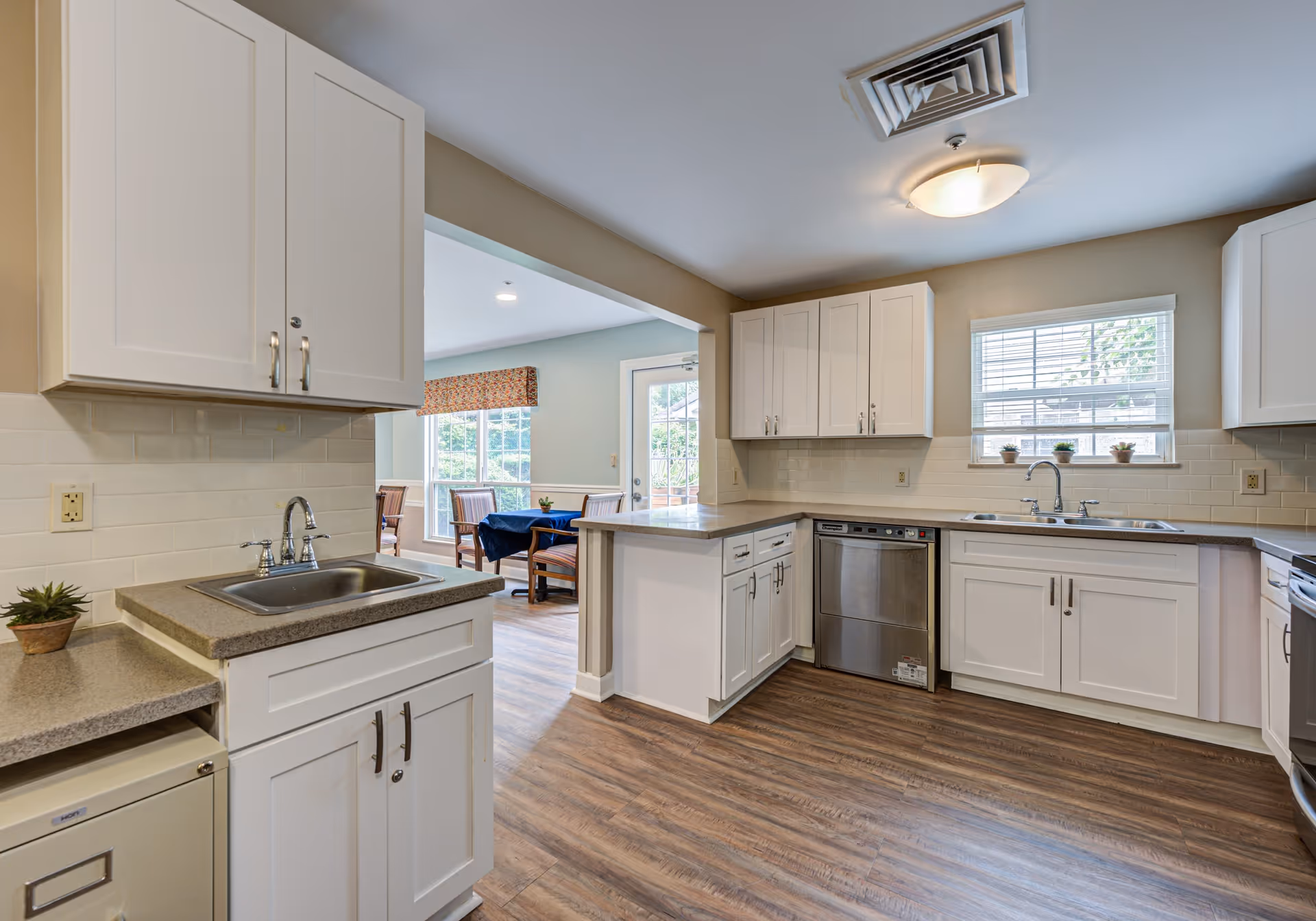 A clean and modern kitchen with white cabinets, a stainless steel dishwasher, two sinks, and a window with small potted plants on the windowsill. The kitchen has wood flooring and opens into a dining area with a table and chairs near large windows.