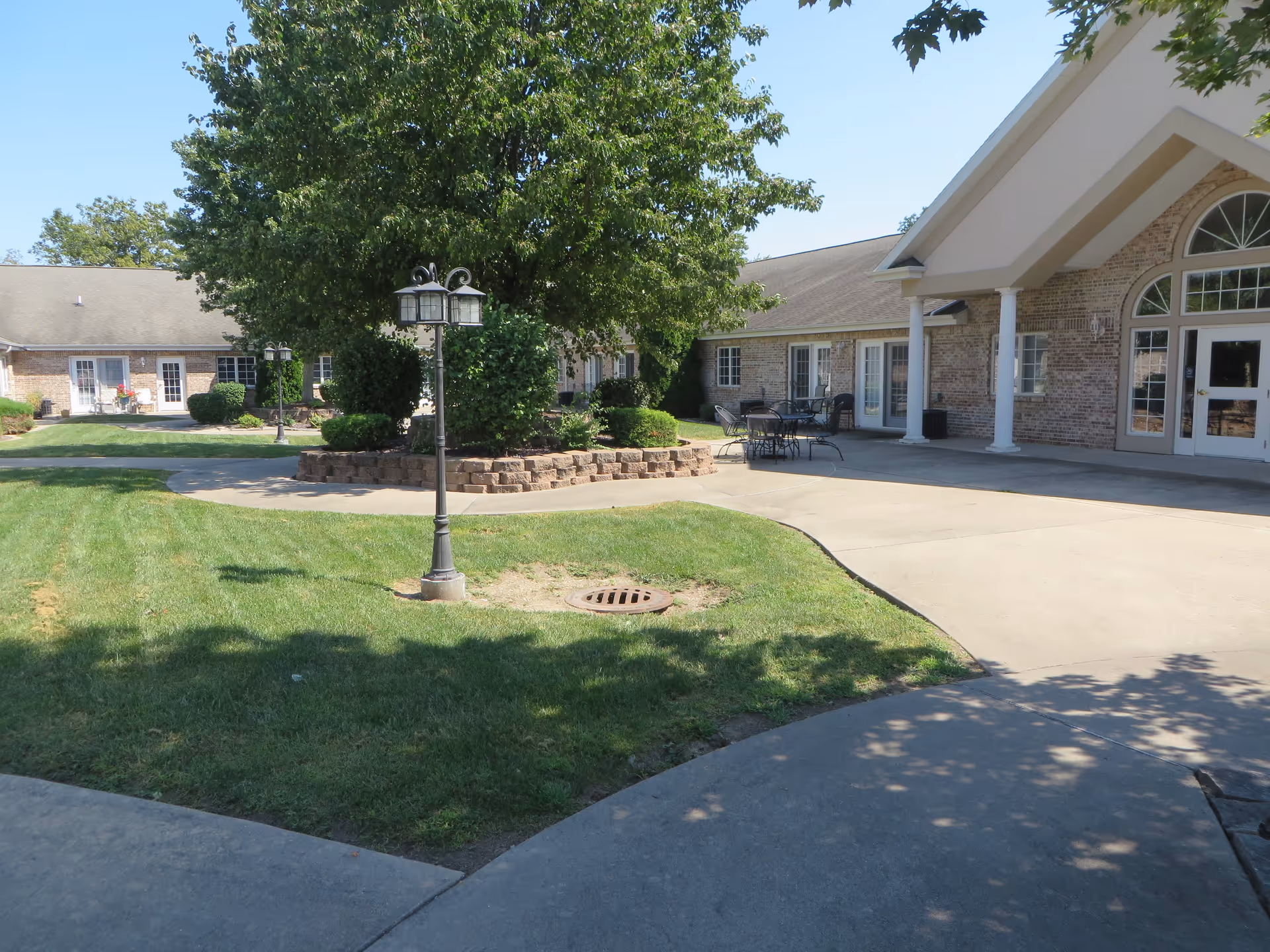 Outdoor courtyard area of a senior living facility with a large tree surrounded by a brick planter, green grass, a lamp post, and patio tables and chairs near the building entrance.
