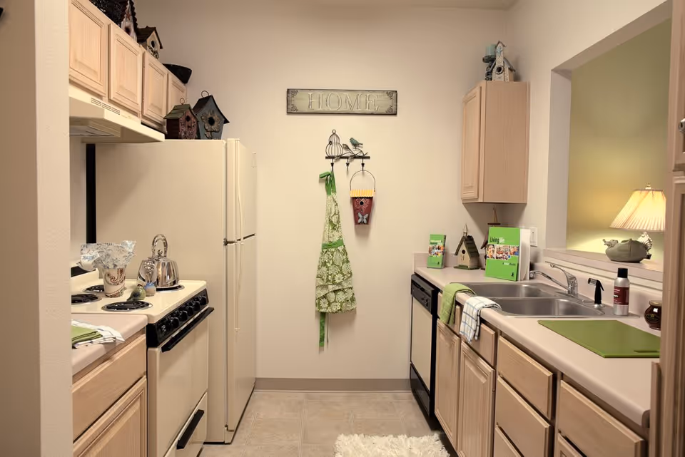 A compact kitchen with light wood cabinets, a white refrigerator, stove, and dishwasher. There is a double sink with a green cutting board on the counter, a green apron hanging on the wall under a decorative 'HOME' sign, and various small decorative items including birdhouses and a lamp visible through a pass-through window.