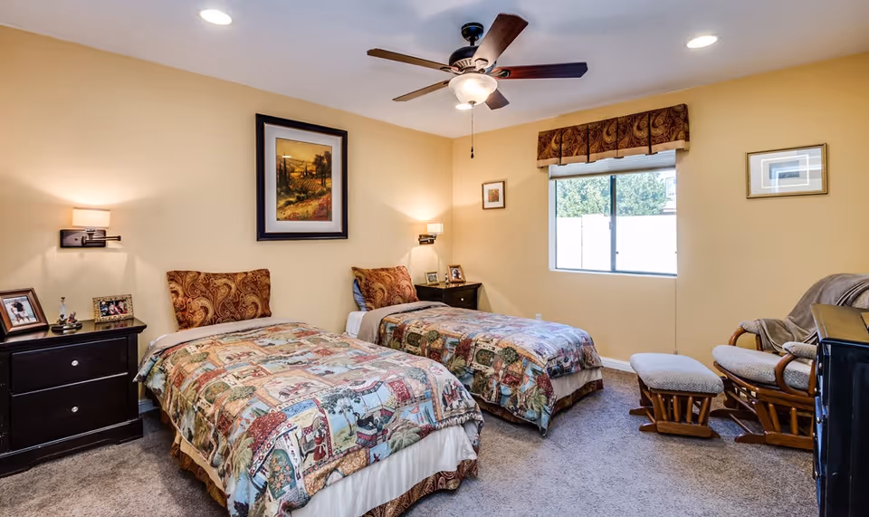A cozy bedroom with two twin beds featuring patterned bedspreads and matching pillows. Each bed has a dark wooden nightstand with framed photos and a wall-mounted lamp above. A ceiling fan with a light hangs from the ceiling. There is a window with a valance letting in natural light, and a cushioned rocking chair with an ottoman is placed near the window. The walls are painted a warm beige color and a framed landscape painting hangs above one bed.