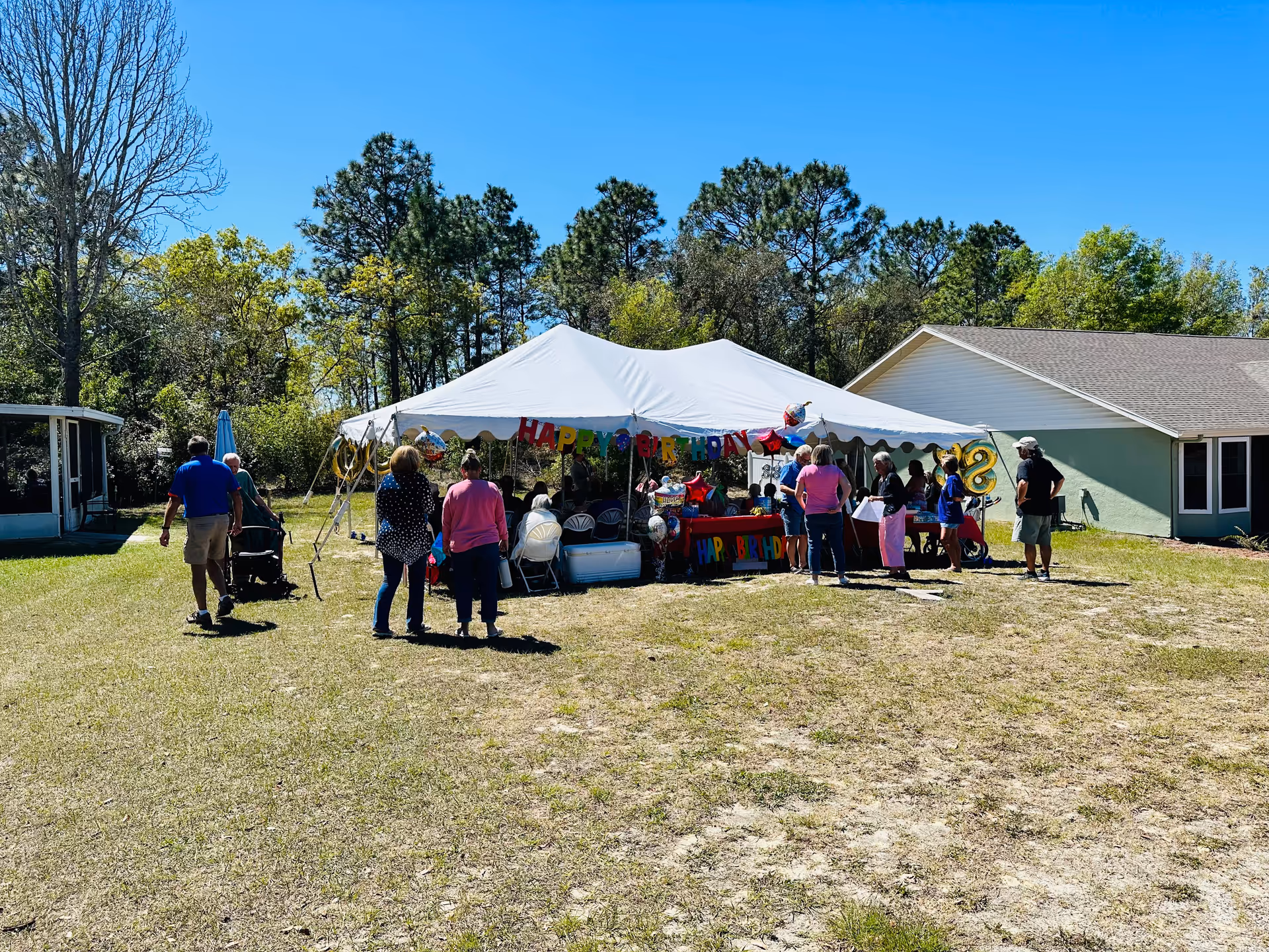 Outdoor birthday celebration at Always There Assisted Living & Memory Care Citrus Springs with a white tent decorated with colorful 'Happy Birthday' banners and balloons. Several people are gathered around and under the tent on a grassy area with trees and buildings in the background under a clear blue sky.