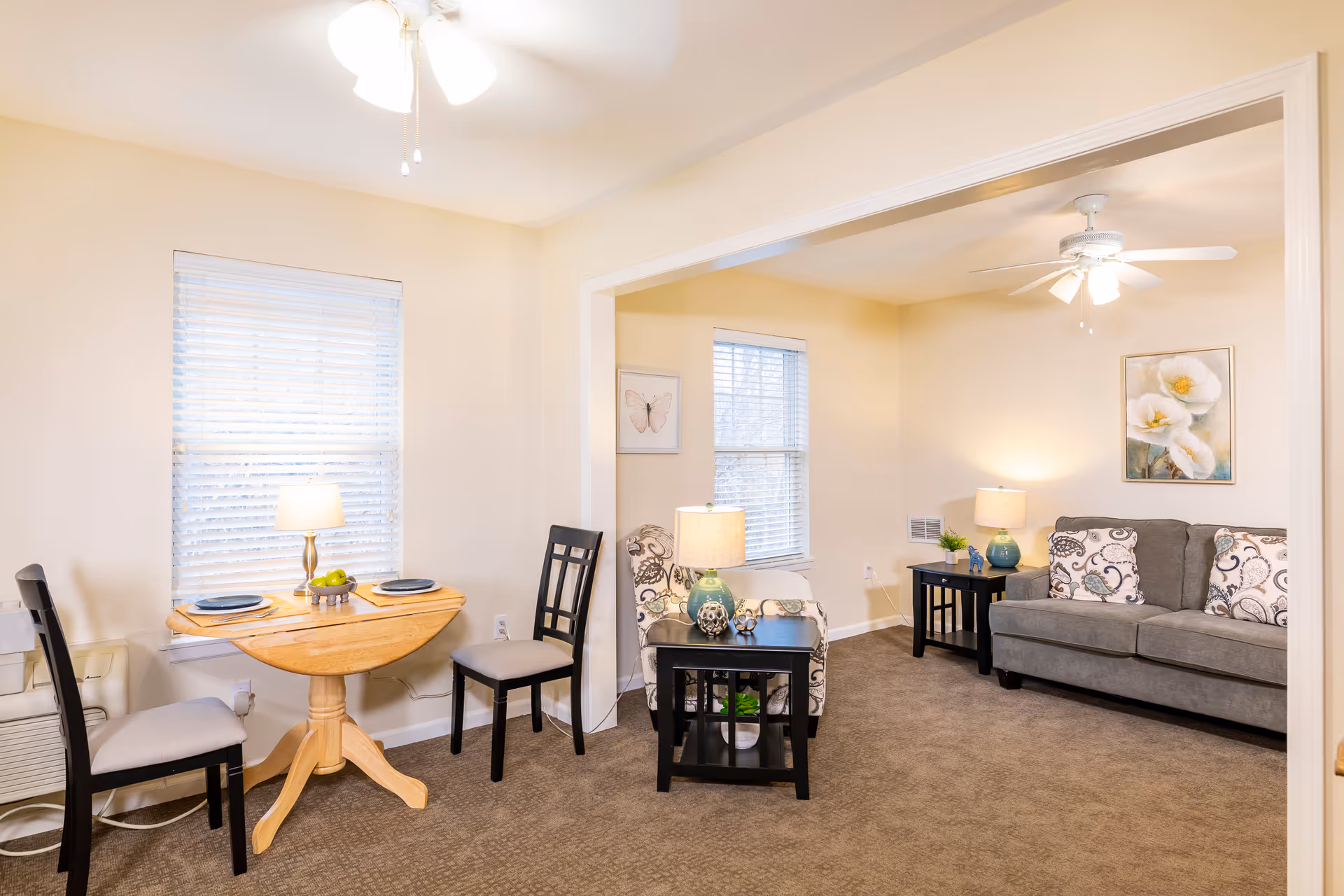 A cozy living and dining area with beige walls and carpeted floor. The dining area features a small wooden round table set for two with black chairs and a table lamp. The living room has a gray sofa with patterned cushions, a patterned armchair, two black side tables with lamps, and framed artwork on the walls. Ceiling fans with lights are installed in both areas.