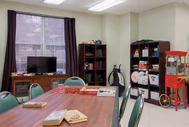 A common room with a large wooden table surrounded by green chairs. On the table are books and board games. In the background, there is a TV on a wooden cabinet, two bookshelves filled with books and games, a golf bag, and a red popcorn machine. The room has beige walls and a window with dark curtains.