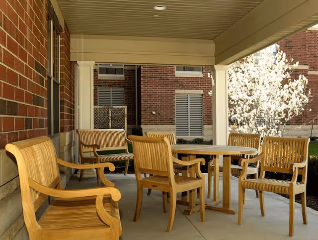 Covered outdoor patio area with wooden chairs and a round wooden table, adjacent to a brick building. A blooming tree is visible in the background.
