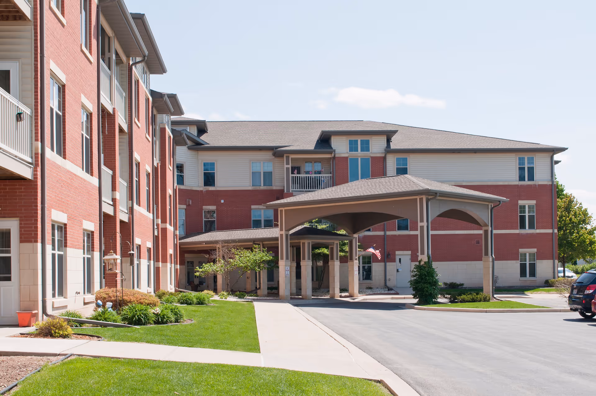 Exterior view of a multi-story senior living facility with red brick and beige siding. The building features multiple windows, balconies, and a covered entrance driveway. There is a well-maintained lawn and some shrubs along the sidewalk leading to the entrance. A few cars are parked near the building under a clear sky.