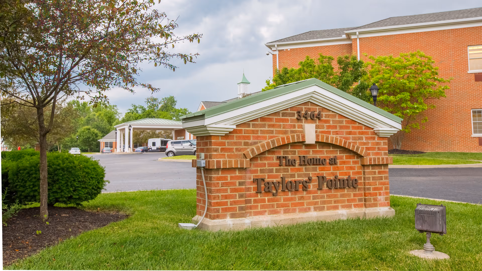 A brick sign with a green roof displaying the text 'The Home at Taylor's Pointe' and the number 3464, situated on a grassy area with trees and bushes nearby. In the background, there is a parking lot with several vehicles and a red brick building under a cloudy sky.