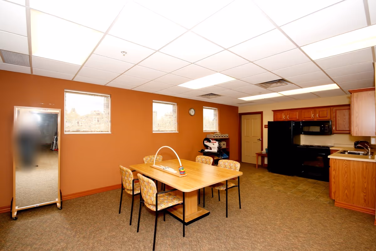 Interior room with a wooden table surrounded by six patterned chairs, a kitchen area with black appliances including a refrigerator, stove, and microwave, wooden cabinets, a sink, and a wall clock above the table. There is a full-length mirror on the left side and three small windows on the orange wall.
