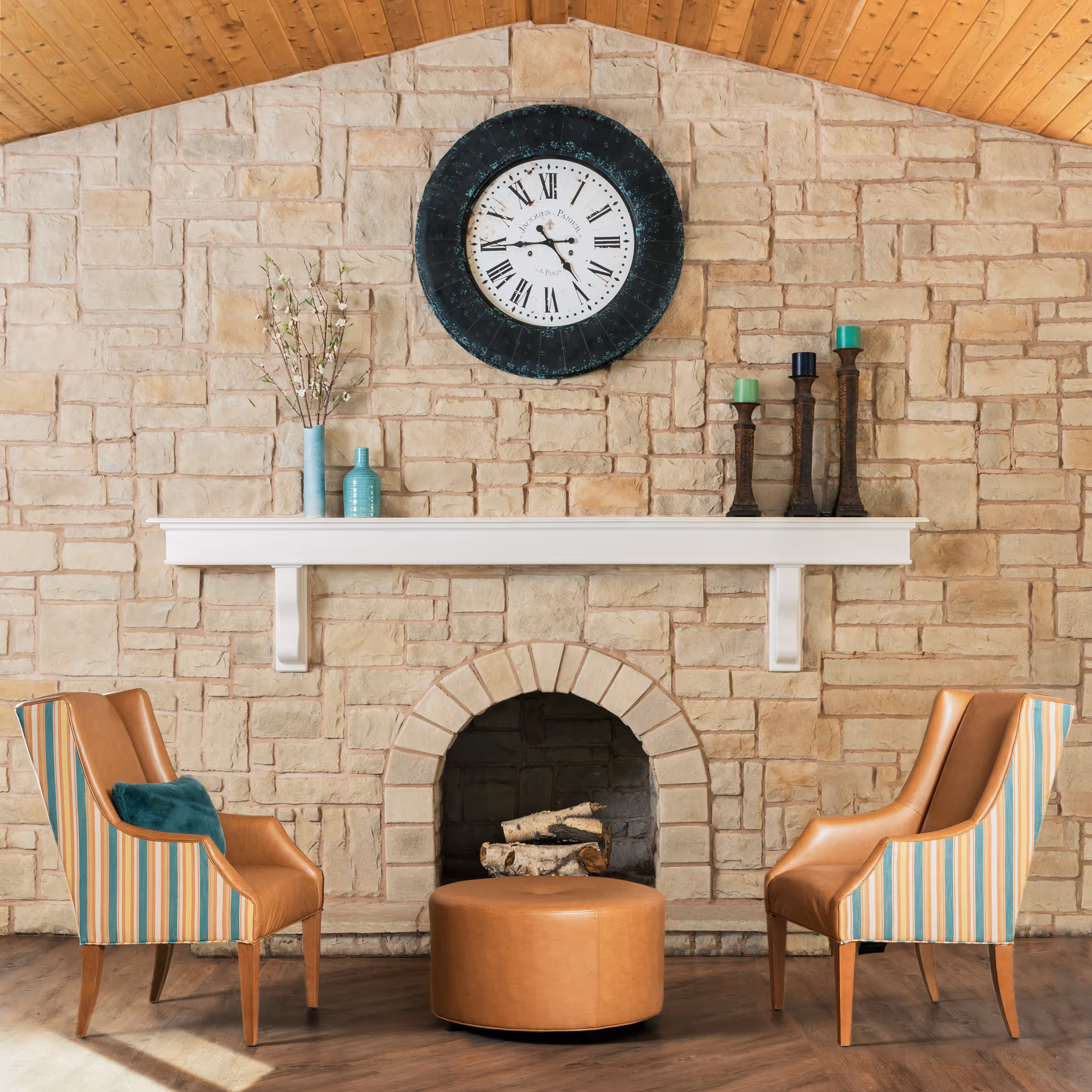 A cozy seating area with two striped and leather armchairs facing each other across a round leather ottoman. Behind them is a stone fireplace with logs inside, a white mantel decorated with vases and candles, and a large round clock with Roman numerals mounted on the stone wall above.