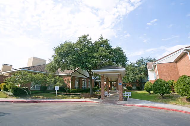 Exterior view of Juniper Village at Lincoln Heights showing a brick building with a covered entrance supported by brick columns, surrounded by green trees and shrubs under a partly cloudy sky.