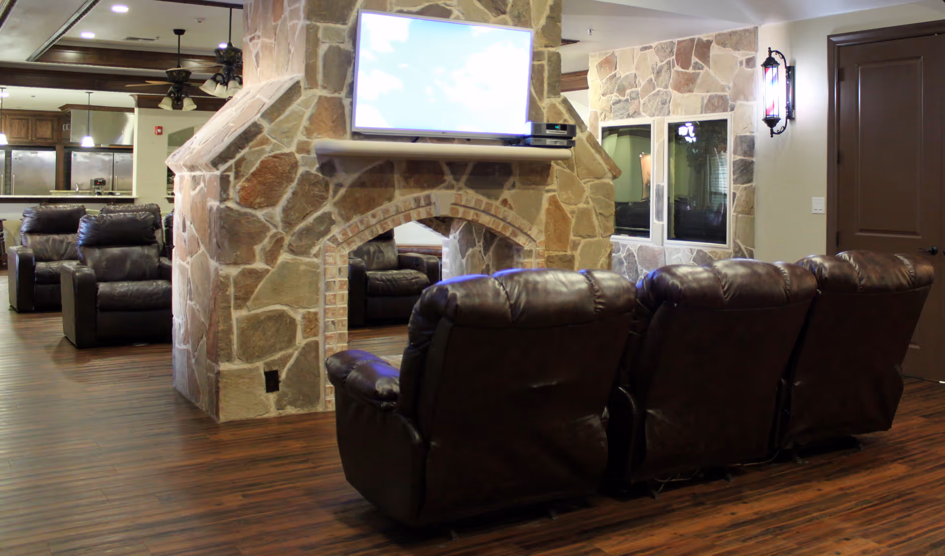 Interior view of a senior living facility lounge area with a stone fireplace in the center. A flat-screen TV is mounted above the fireplace. There are several dark brown leather recliners arranged facing the TV. The room has wooden flooring and a kitchen area is visible in the background.