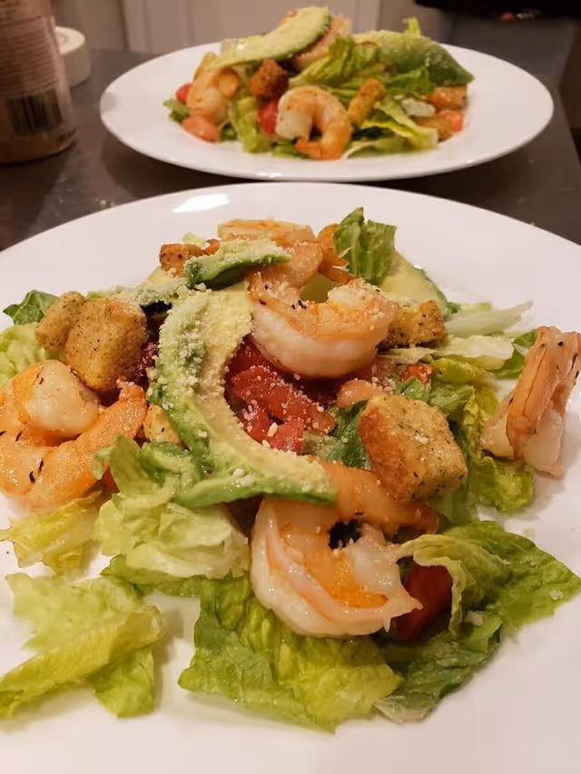 Two plates of shrimp salad with lettuce, avocado slices, croutons, diced tomatoes, and grated cheese on a kitchen counter.