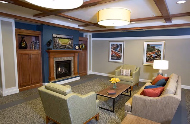 A cozy living room with a beige sofa adorned with red and blue pillows, two green armchairs, a wooden coffee table with a vase of yellow flowers, a fireplace with a wooden mantel, built-in wooden cabinets on either side, and framed artwork on the walls under a ceiling with wooden beams and a large round light fixture.