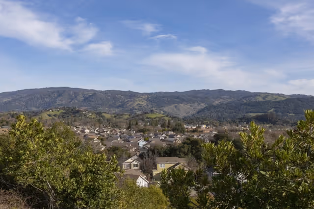 View of a residential neighborhood with houses surrounded by trees and greenery, set against a backdrop of rolling hills under a partly cloudy blue sky.