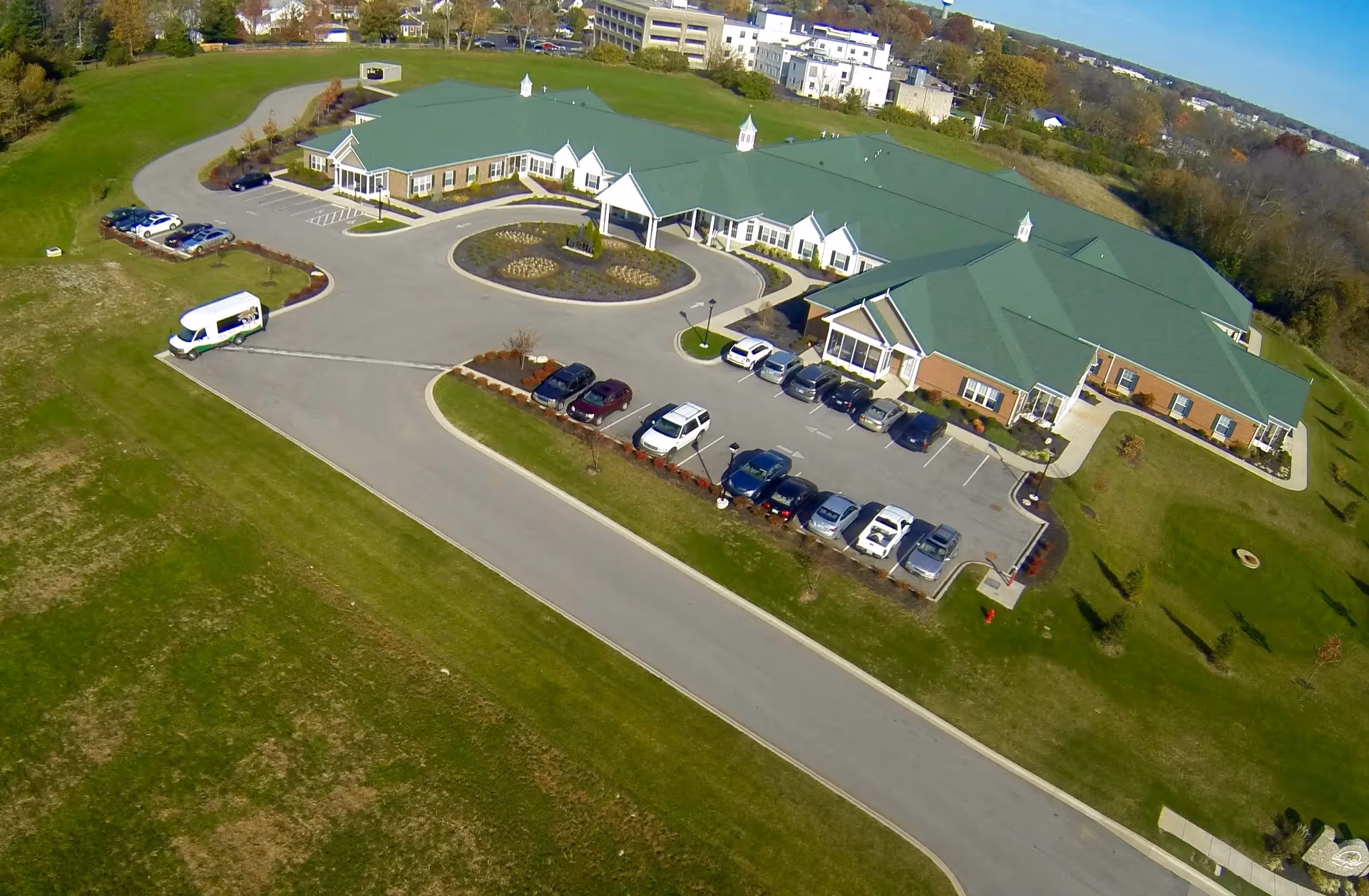 Aerial view of Daisy Hill Senior Living facility showing a large single-story building with green roofs, surrounded by parking lots with several cars, landscaped areas, and green lawns under a clear blue sky.