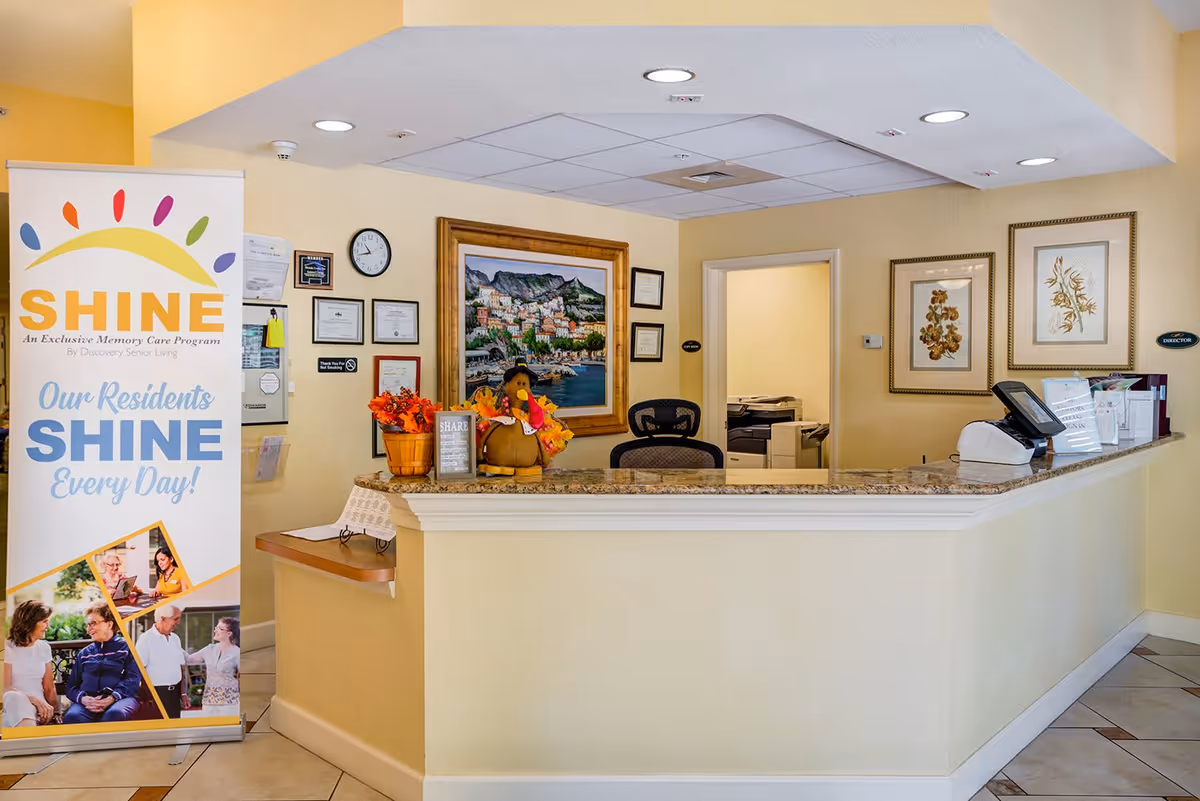 Reception desk area in a senior living facility with a granite countertop, office chair, and decorative items including a stuffed turkey and a basket with autumn leaves. On the left side, there is a standing banner promoting the SHINE memory care program. The walls are decorated with framed artwork and certificates.