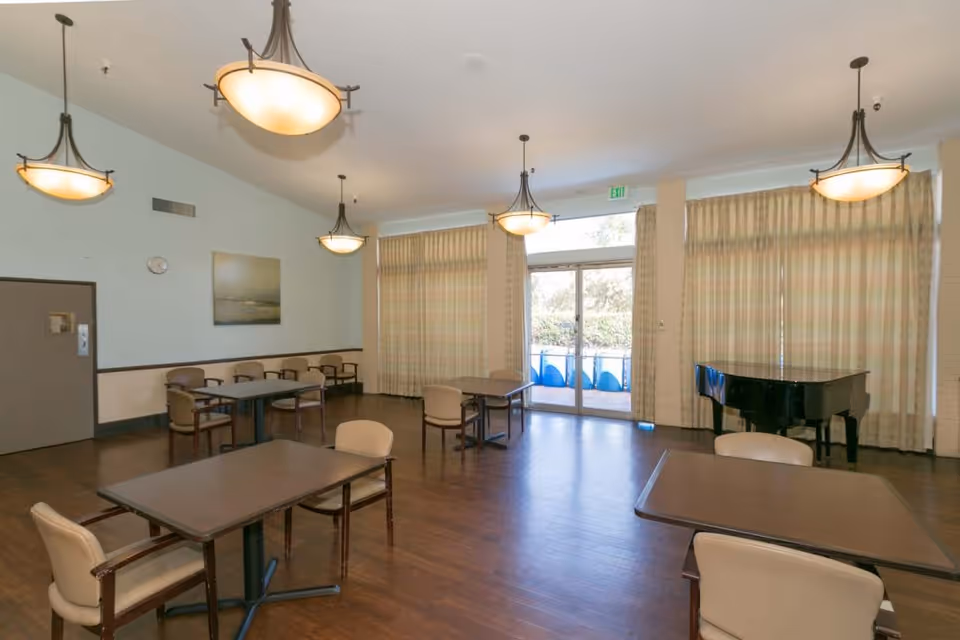 A spacious room with several tables and chairs arranged for seating, large windows with curtains allowing natural light, a black grand piano in the corner, and multiple hanging ceiling lights providing illumination.