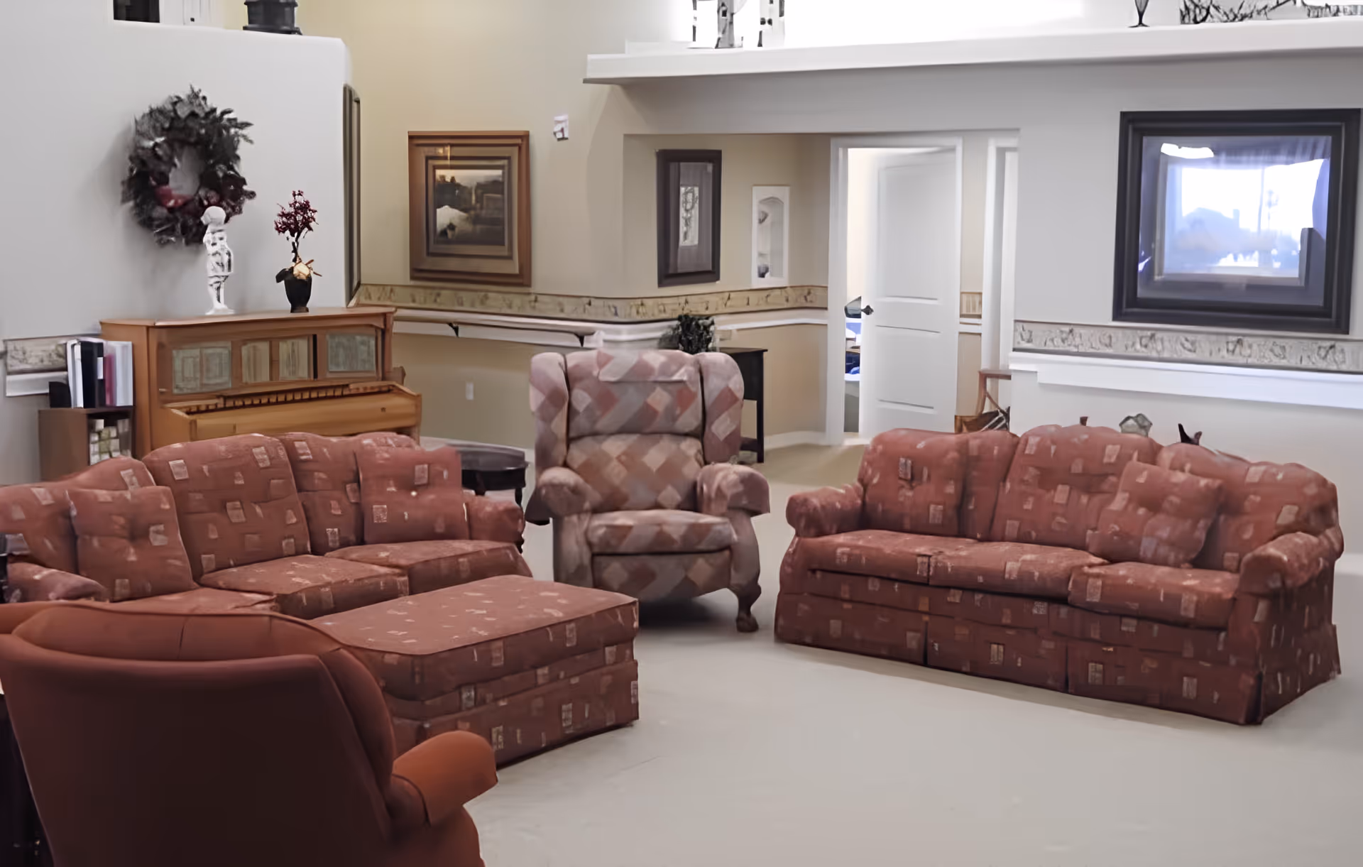 A cozy living room area with two patterned red sofas, a matching armchair, and a cushioned ottoman. There is a piano against the wall decorated with a wreath and a small statue. The walls are light-colored with framed artwork and a window. The room has a soft carpet and an open doorway leading to another room.