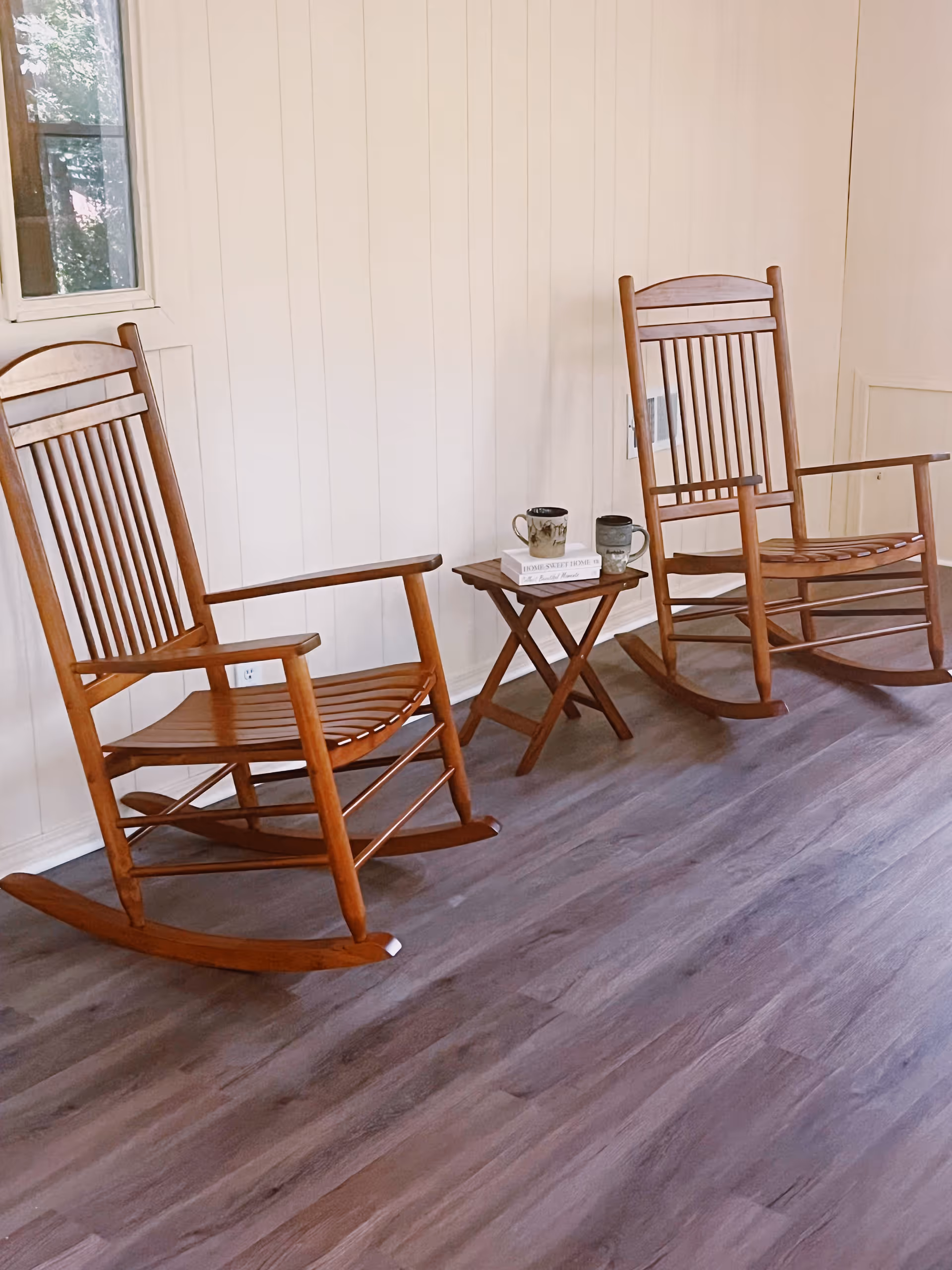 Two wooden rocking chairs with slatted backs and seats are positioned on a wood floor against a white paneled wall. Between the chairs is a small wooden folding table holding two coffee mugs and a book titled 'Home Sweet Home'. A window is visible on the left side of the wall.