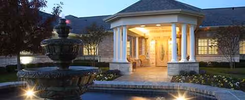 Front entrance of Grand Brook Memory Care of McKinney with lit portico, columns, and fountain.