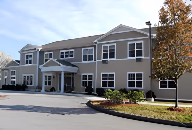 Exterior view of a two-story senior living facility building with multiple windows, a covered entrance with columns, a tree with autumn leaves, and a paved driveway in front.