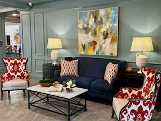 A cozy living room area with a dark blue sofa adorned with patterned pillows, two red and white patterned armchairs, a rectangular coffee table with decorative items, two table lamps on side tables, and a colorful abstract painting on the wall.