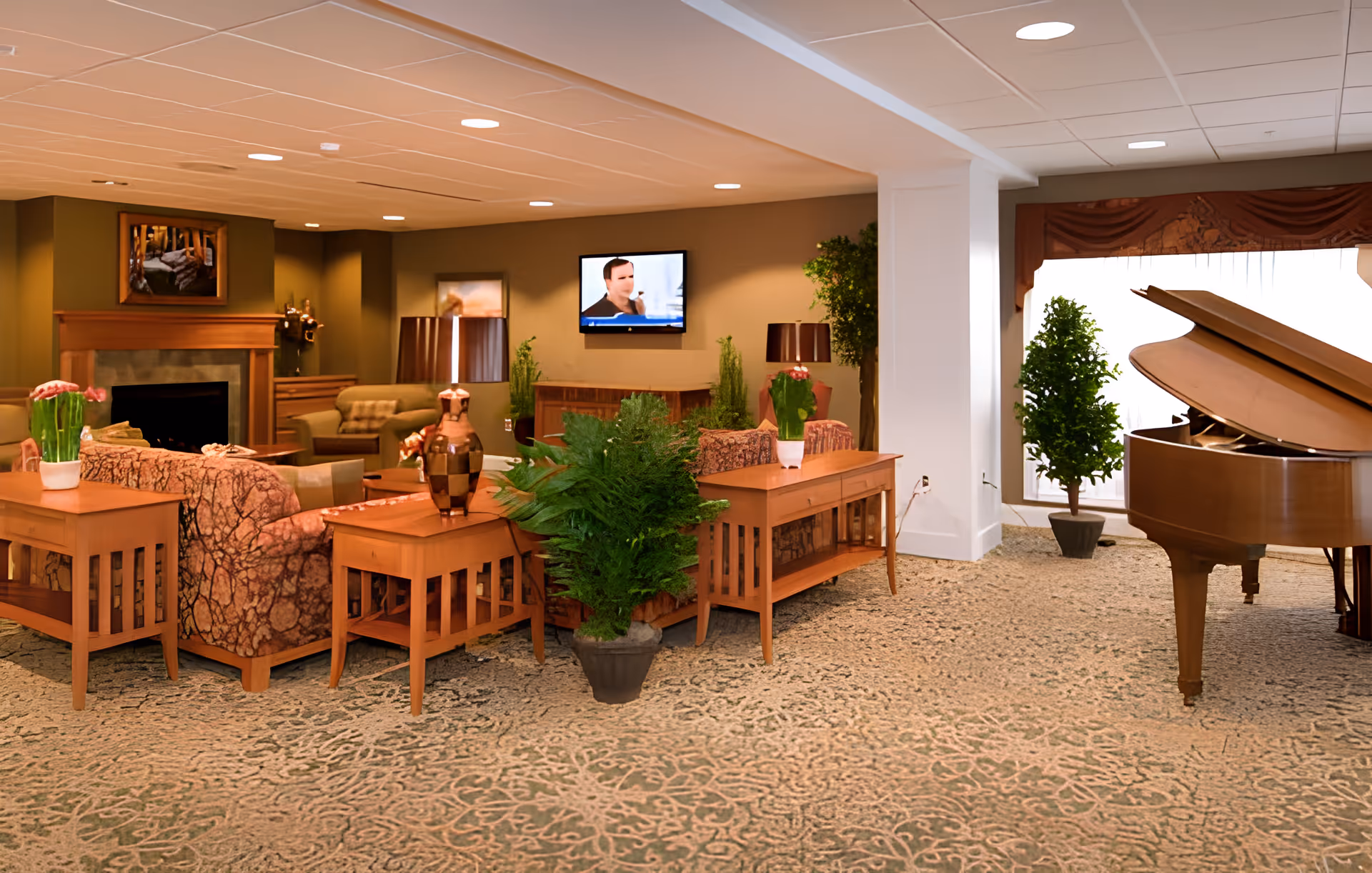 Spacious assisted living lounge with sofas, wooden tables, plants, a fireplace, a wall-mounted TV and a grand piano.