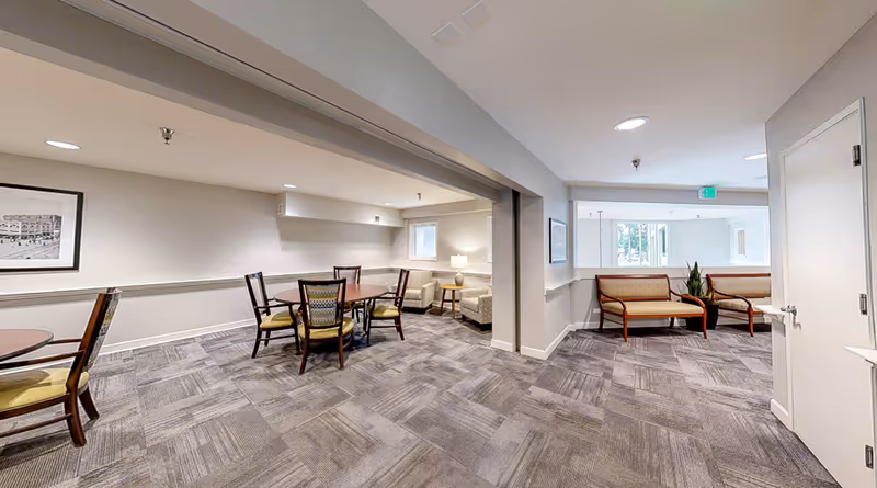 Spacious assisted-living common area with round tables, chairs, and bench seating under recessed lighting.