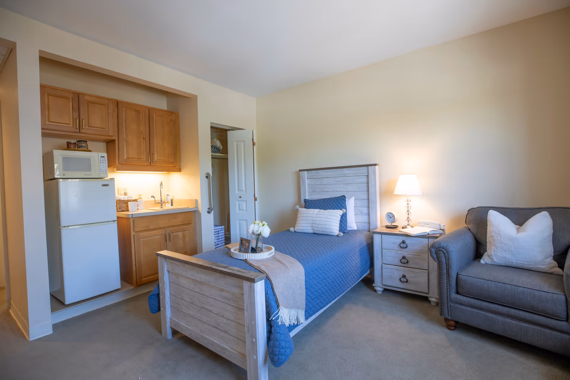 A cozy bedroom in a senior living facility featuring a single bed with blue bedding and decorative pillows, a wooden nightstand with a lamp, clock, and telephone, a gray armchair with a white pillow, and a small kitchenette area with a refrigerator, microwave, sink, and wooden cabinets.