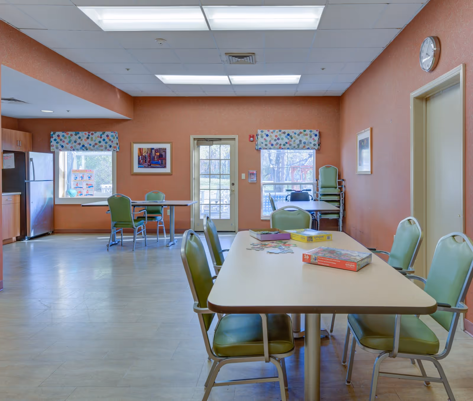A brightly lit room with orange walls featuring several tables and green chairs arranged around them. On the tables are puzzle boxes and partially completed puzzles. There are two windows with colorful polka dot valances, a door leading outside, a clock on the wall, and a refrigerator in the corner.