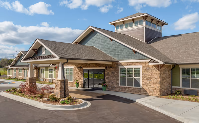 Exterior front view of a single-story senior living facility building with stone and siding facade, large windows, a covered entrance supported by stone pillars, and a tower-like structure on the roof under a partly cloudy sky.