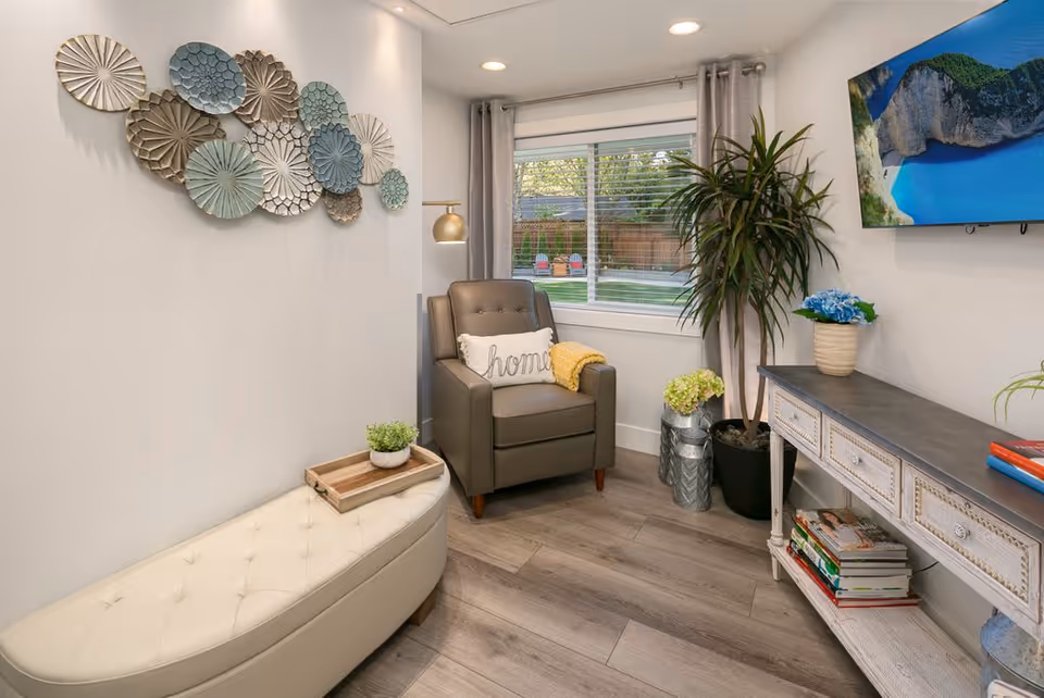 A cozy living room corner with a gray armchair featuring a white pillow with the word 'home' and a yellow throw blanket. Next to the chair is a small white tufted bench with a wooden tray and a small potted plant. The wall has decorative metal wall art in various circular patterns. A large window with gray curtains shows a view of a backyard with colorful chairs. A tall potted plant and a white console table with books and a vase with blue flowers are also visible. A flat-screen TV is mounted on the wall above the console table.