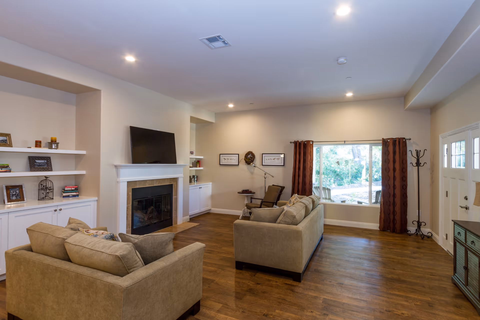 A spacious living room with two beige sofas facing a fireplace with a mounted flat-screen TV above it. The room has wooden floors, built-in white shelves with decorative items, a large window with brown curtains, a coat rack, and a door with glass panels.