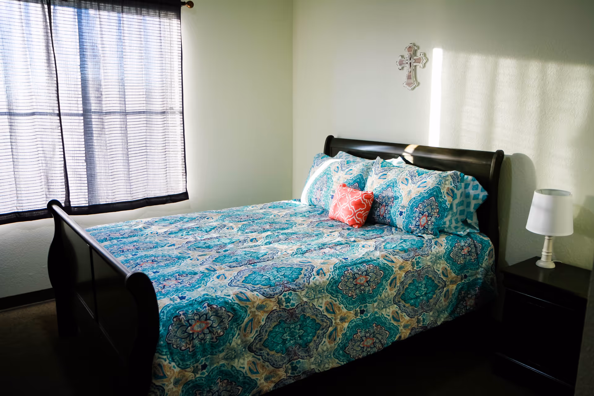 Bright bedroom featuring a dark wood sleigh bed dressed in a teal patterned comforter with a red accent pillow, a nightstand and lamp, and a curtained window.
