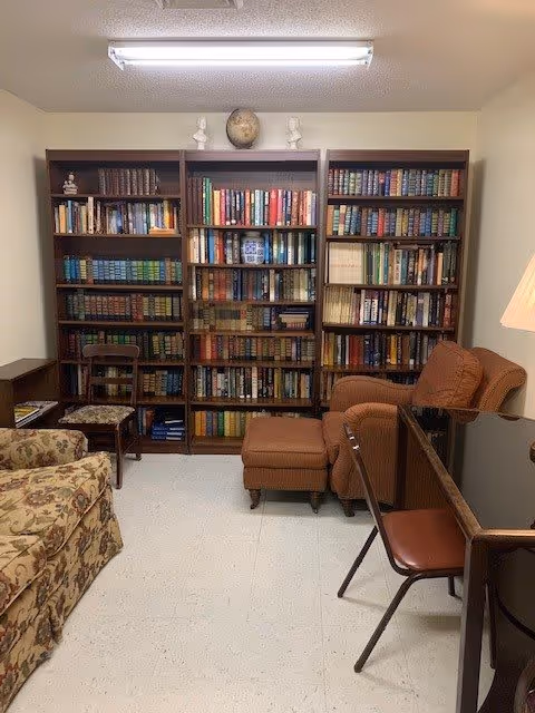 A cozy reading room with three large wooden bookshelves filled with books. There is a floral patterned armchair on the left, a wooden chair with a cushioned seat, and a brown upholstered armchair with a matching ottoman. A wooden table with a brown chair is on the right side, and a lamp is partially visible. The room has a white tiled floor and a fluorescent ceiling light.