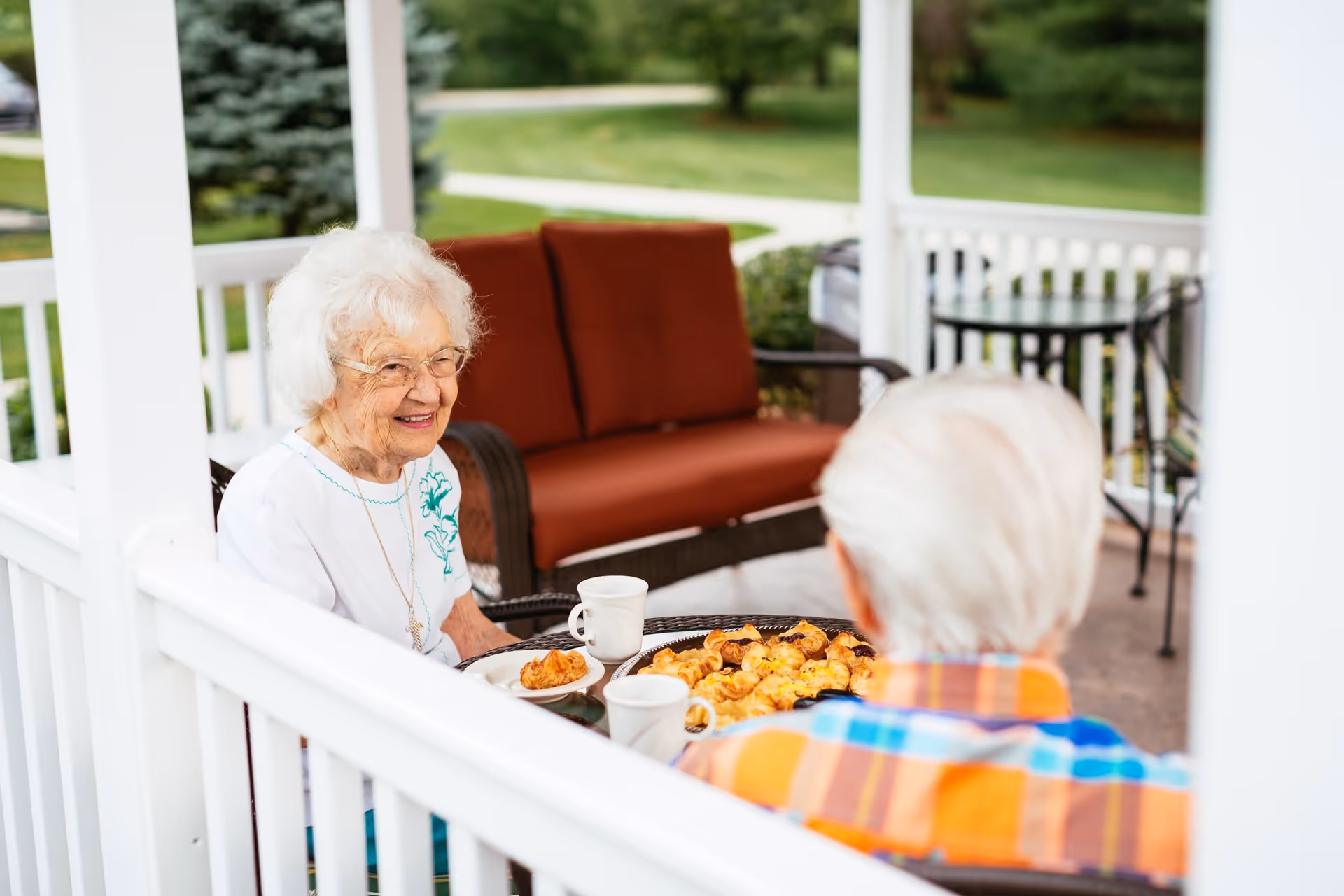 Two elderly individuals sitting at a round table on a covered outdoor porch, enjoying pastries and coffee. One person is smiling and wearing glasses and a white shirt with a floral design, while the other is seen from behind wearing a colorful plaid shirt. The porch has white railings and a cushioned bench with rust-colored cushions in the background, with green grass and trees visible beyond the porch.