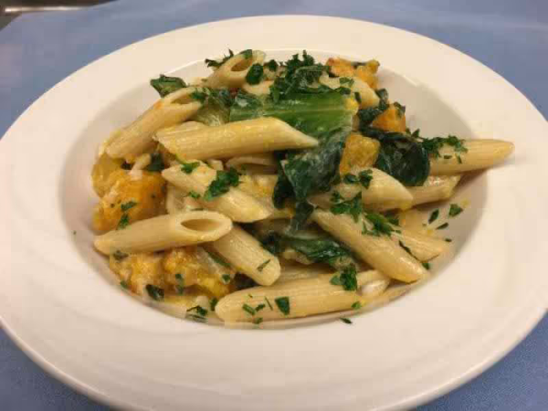 A white bowl filled with penne pasta mixed with leafy greens and chunks of yellow squash, garnished with chopped herbs, placed on a blue tablecloth.