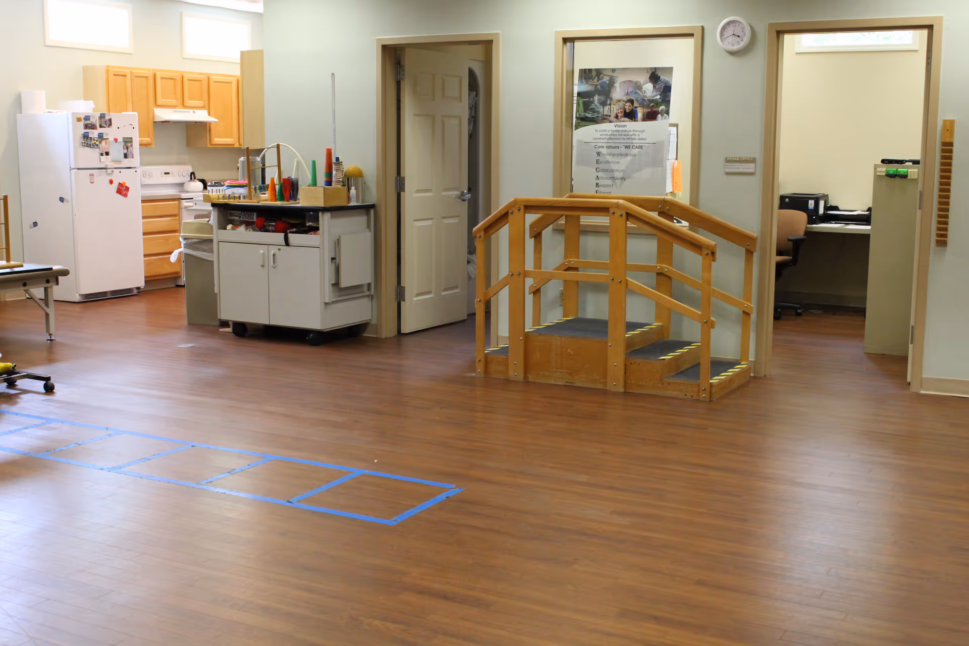Interior view of a room in a senior living facility with wooden flooring, a small wooden staircase with handrails, a kitchenette area with a refrigerator, stove, and cabinets, and an open doorway leading to an office space with a desk and chair.