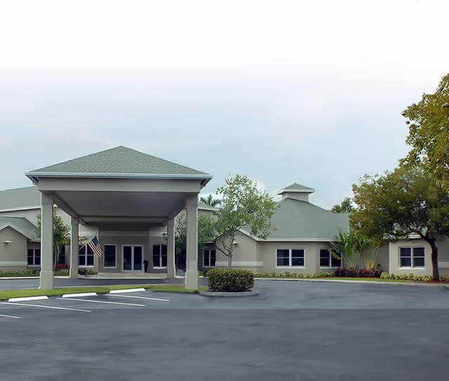 Exterior view of a single-story building with a covered entrance supported by columns. The building has a green roof and beige walls, surrounded by trees and shrubs. There is an American flag hanging near the entrance and an empty parking lot in the foreground.