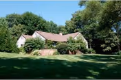 A single-story house with a light-colored exterior and a brown roof, surrounded by lush green trees and a well-maintained grassy lawn under a clear sky.