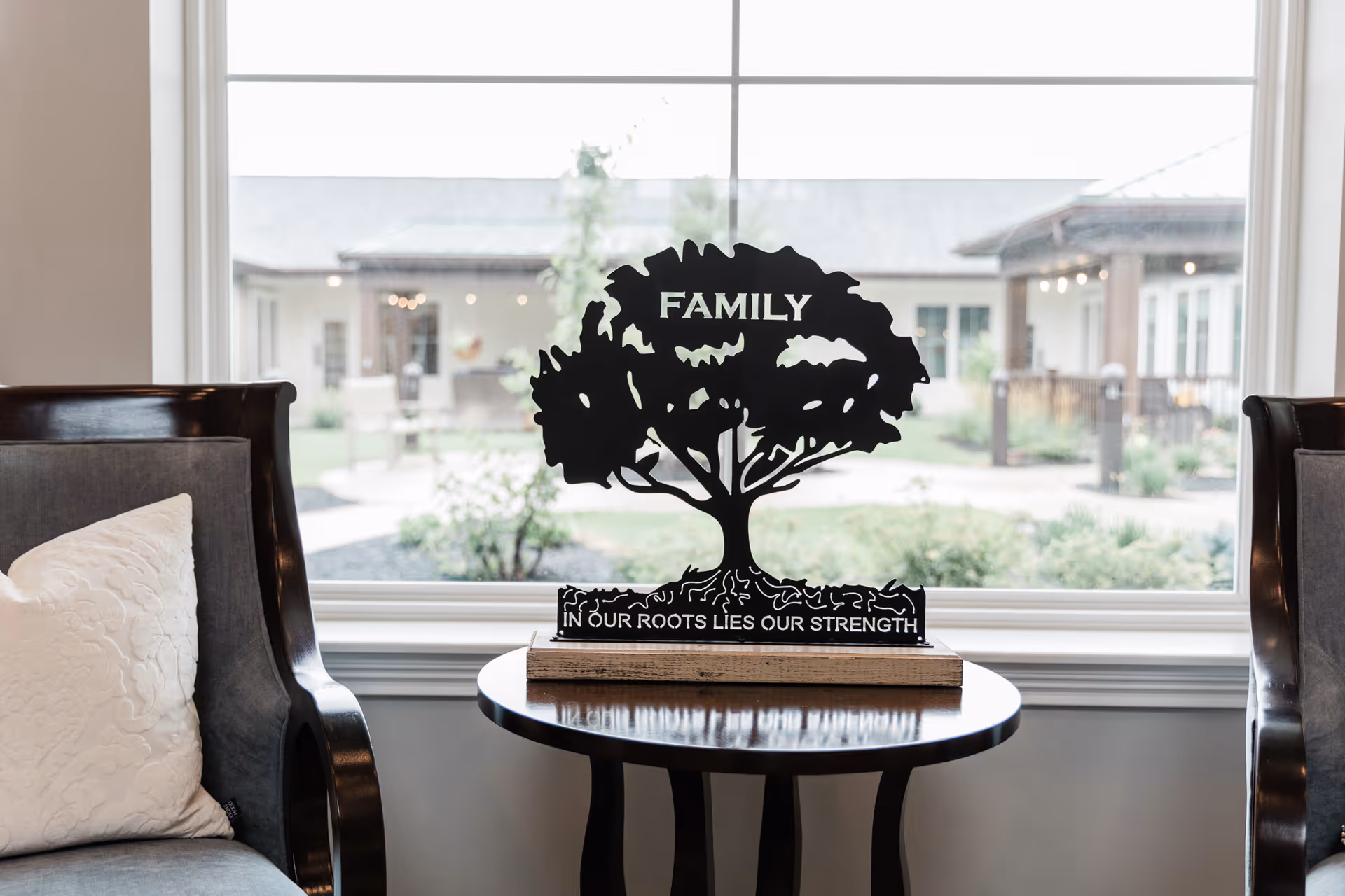 A decorative black metal tree sculpture with the word 'FAMILY' on the tree and the phrase 'IN OUR ROOTS LIES OUR STRENGTH' on the base, placed on a round wooden table between two dark wooden chairs with gray cushions and a white pillow. A large window behind the table shows an outdoor view of a building and greenery.