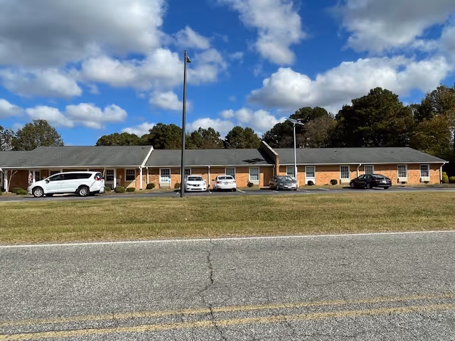 Single-story brick building with a gray shingled roof, several windows, and a few parked cars in front under a partly cloudy blue sky.