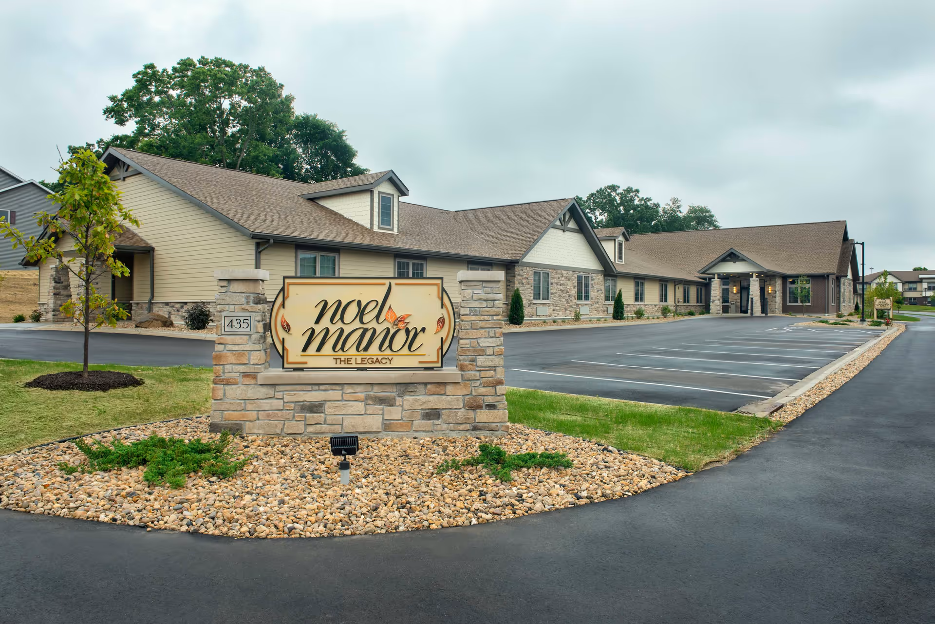 Stone entrance sign reading 'Noel Manor' in front of a single-story senior living building with a paved parking lot.