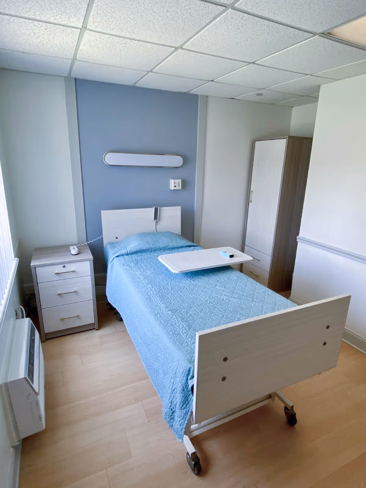 A small, simple bedroom in a care facility with a single hospital-style bed covered with a light blue quilted blanket. The bed has wheels and a white overbed table resting on it. Next to the bed is a small bedside table with three drawers and a call button on top. There is a white and wood wardrobe in the corner, light wood flooring, and a blue accent wall behind the bed. The ceiling has white acoustic tiles and a fluorescent light fixture.
