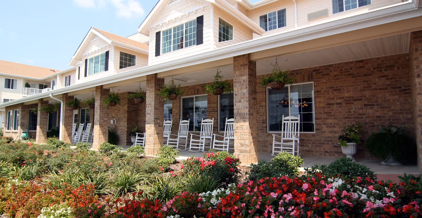 Exterior view of a senior living facility with a covered porch featuring white rocking chairs and hanging plants. The building has brick walls and multiple windows, with a well-maintained garden full of colorful flowers in the foreground.