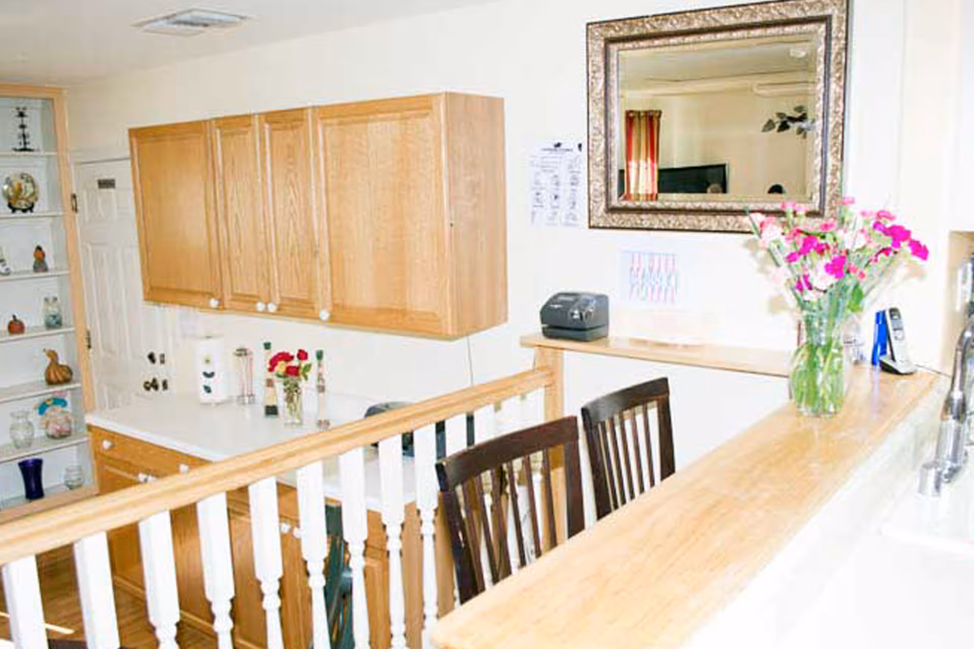 Interior view of a kitchen area with light wood cabinets, a white countertop, a wooden railing with white spindles, two dark wooden chairs, a large ornate mirror on the wall, a vase with pink and white flowers on the counter, and various small decorative items on shelves and counters.