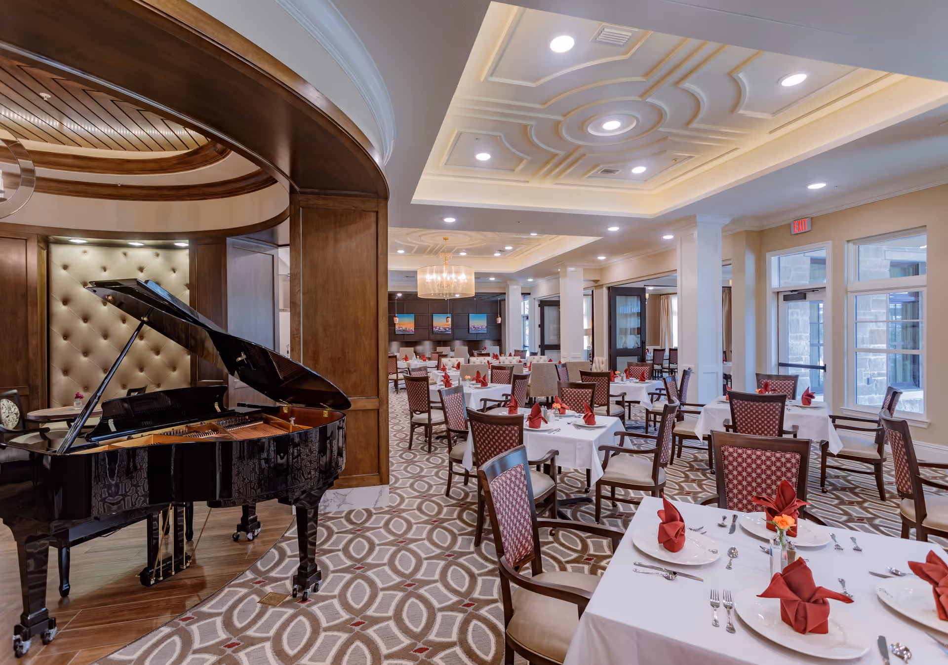 Elegant dining room with multiple tables covered in white tablecloths, each set with plates, silverware, and red folded napkins. A black grand piano is positioned under a wooden archway on the left side. The room features patterned carpet, large windows, and a chandelier hanging from a decorative ceiling.
